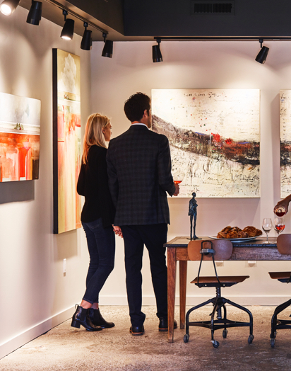 Two people standing, looking at a painting, next to a table set with food and drinks.